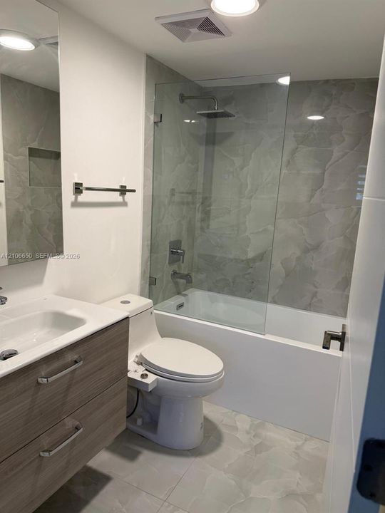 Bathroom featuring a wood-finish vanity with integrated sink, glass-enclosed shower, rainfall showerhead, and large-format neutral tile flooring