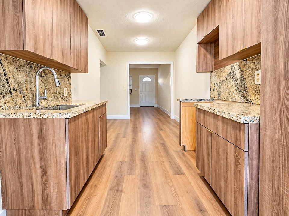 Kitchen featuring wood-finish cabinetry, stone-surface countertops, stone backsplashes, a stainless steel under-mount sink with a high-arch faucet, and wood-finish flooring