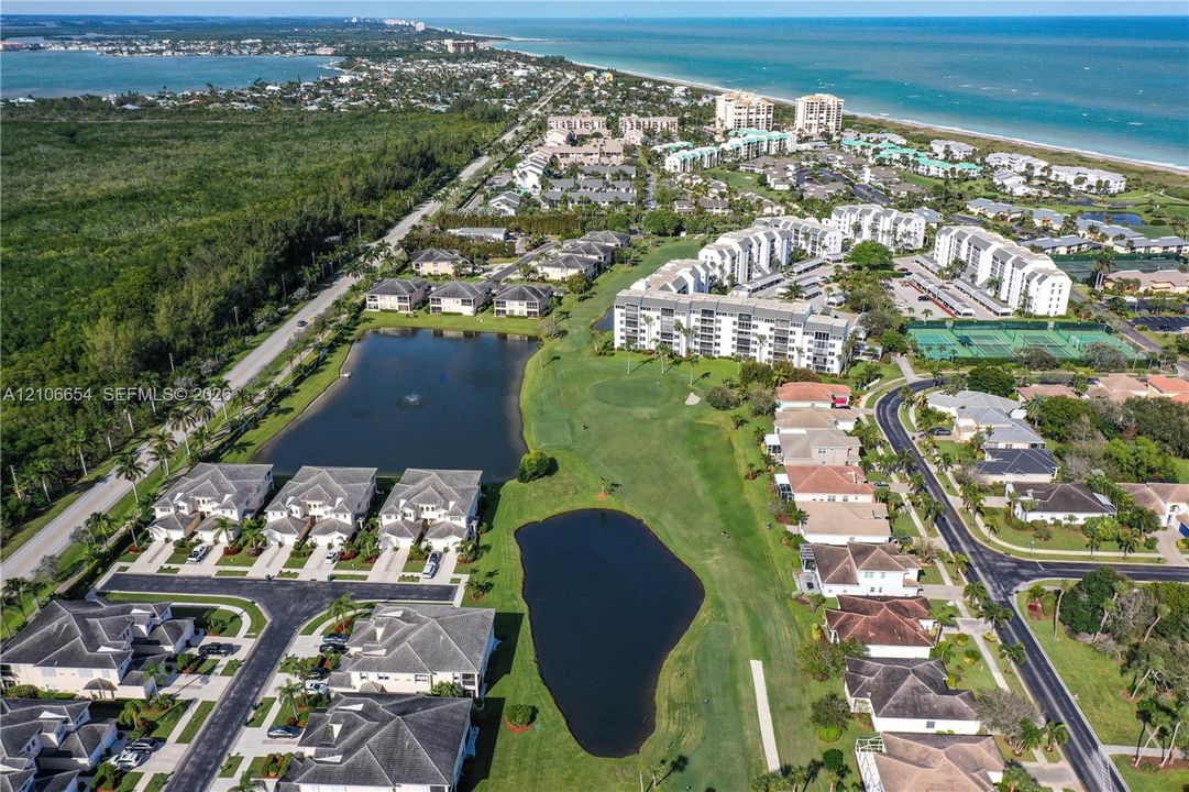 Aerial perspective showcasing a coastal community featuring multi-story residential buildings, manicured golf course grounds, multiple retention ponds, and proximity to the shoreline