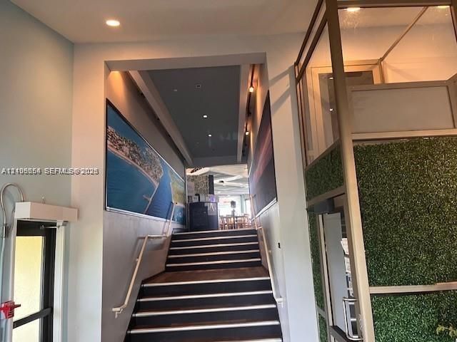 Staircase featuring dark tread flooring, white metal handrails, recessed ceiling lighting, and an enclosed glass elevator with integrated faux-foliage paneling