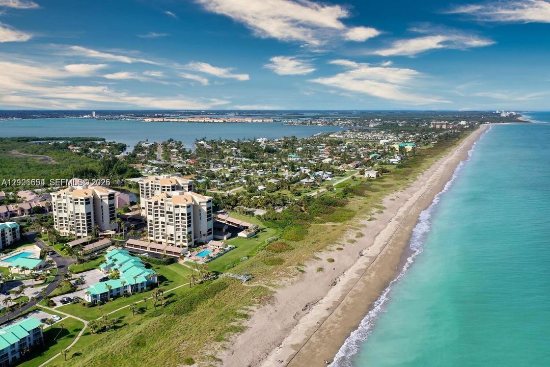 Aerial perspective featuring a multi-story residential complex situated directly along the coastline, proximity to the ocean, expansive sandy beach, and landscaped grounds