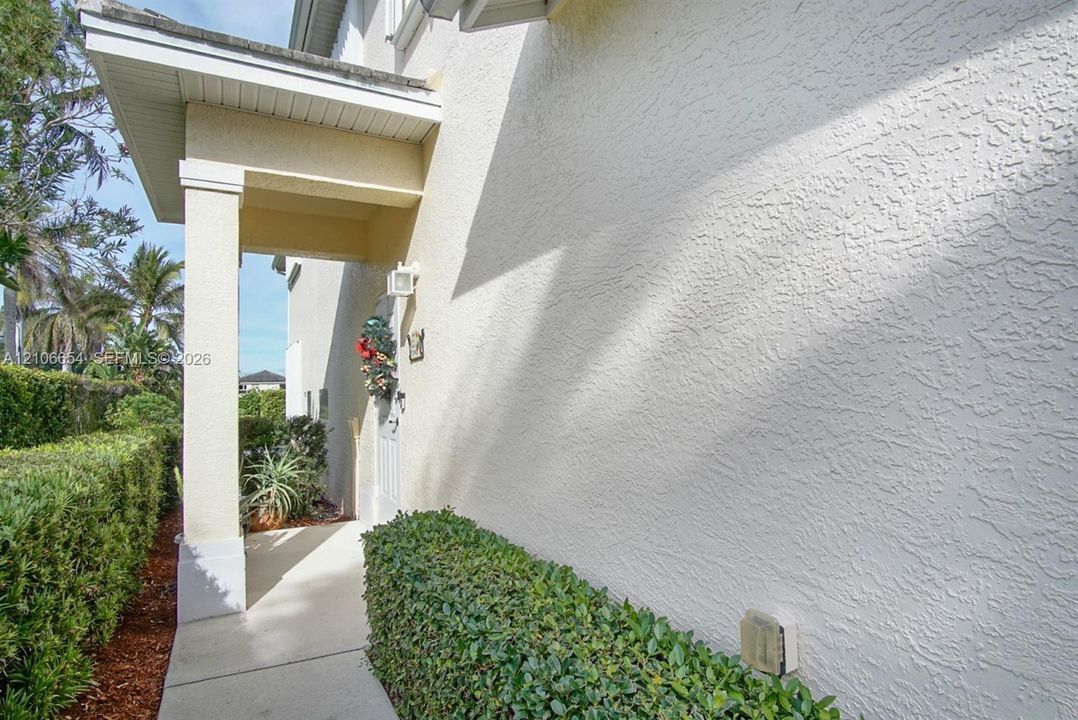 Exterior entry featuring a textured stucco facade, covered porch, square structural column, and concrete walkway bordered by manicured shrubs