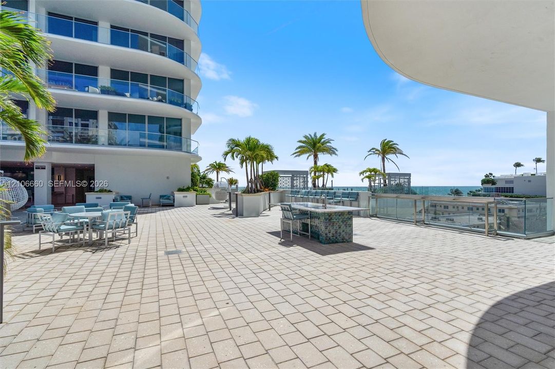 Expansive paved terrace featuring a tiled outdoor kitchen island, glass-panel perimeter railing, and mature palm landscaping