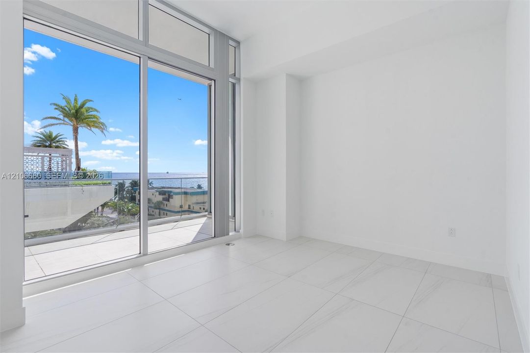 Floor-to-ceiling glass wall, large-format tile flooring, and sliding glass door opening to a private balcony with glass railing and water backdrop
