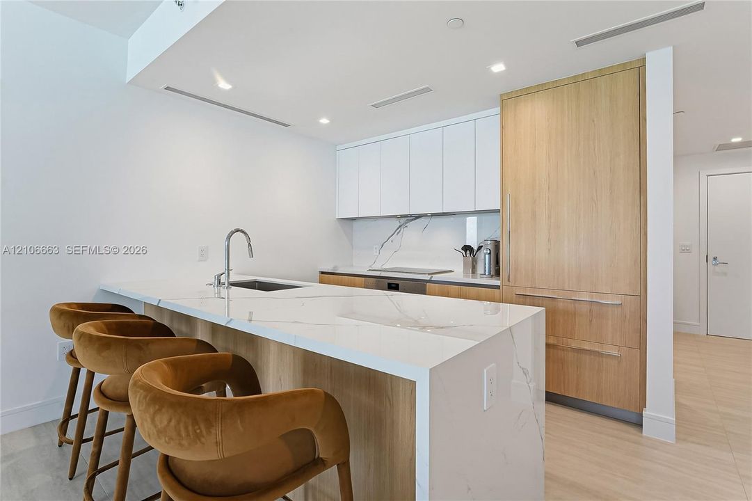 Kitchen featuring waterfall-edge stone countertops, custom cabinetry with wood-finish paneling, integrated refrigerator, stone backsplash, and stainless-steel sink with a gooseneck faucet