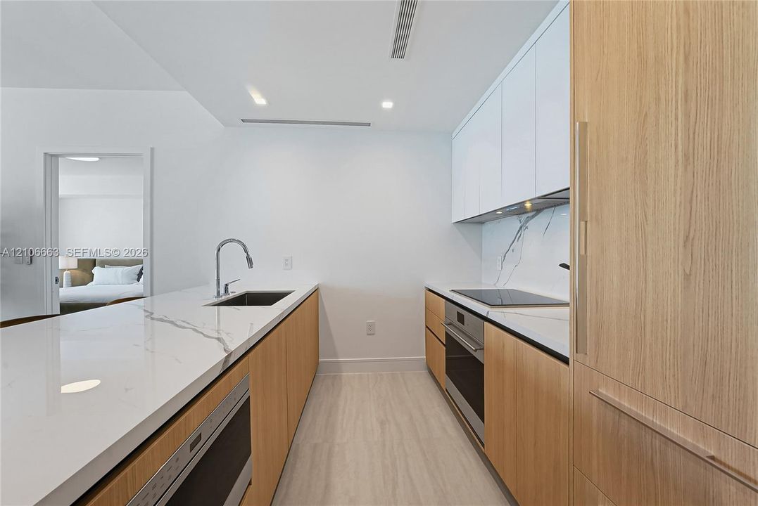 Contemporary kitchen featuring white flat-panel upper cabinetry, light wood-finish lower cabinetry, stone-style countertops with integrated backsplash, built-in cooktop, and undermount stainless steel sink with gooseneck faucet