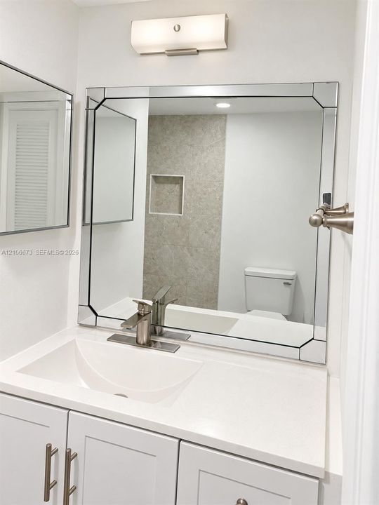 White vanity with a solid-surface countertop and integrated sink, framed beveled wall mirror, brushed-nickel faucet, satin-finish wall lighting, and tiled wall niche