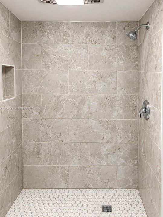 Walk-in shower featuring large-format neutral stone-finish wall tile, white hexagonal floor tile, chrome shower hardware, and a built-in recessed wall niche