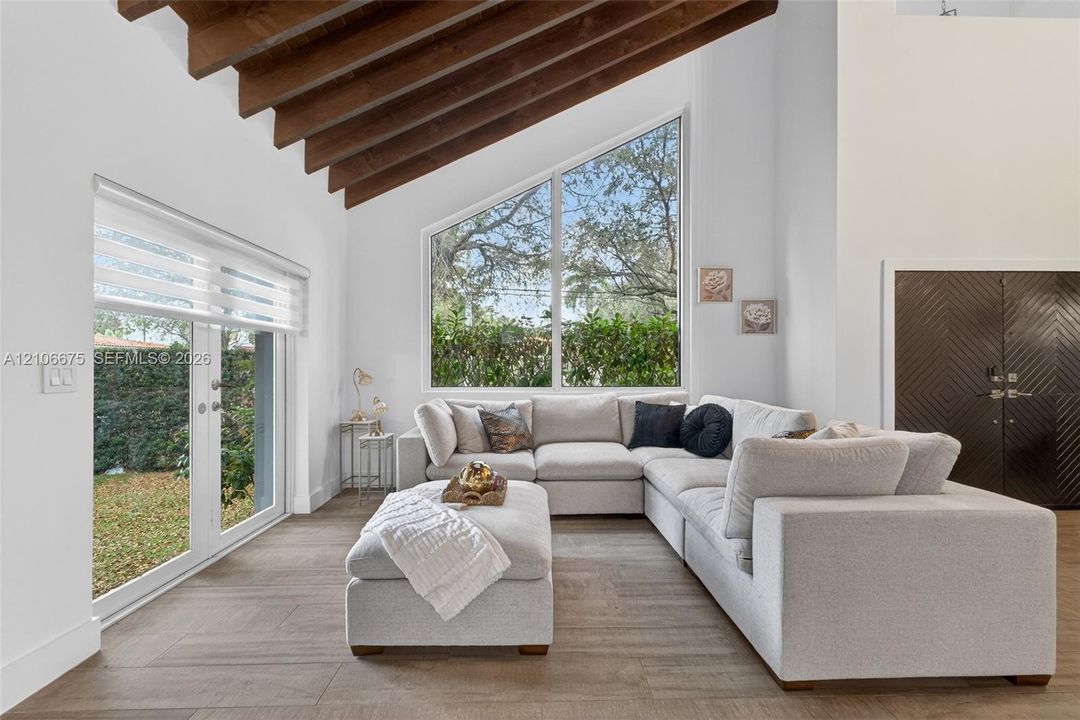 Vaulted ceilings with exposed wood-finish beams, large trapezoidal window, French doors with integrated blinds, and wood-finish tile flooring