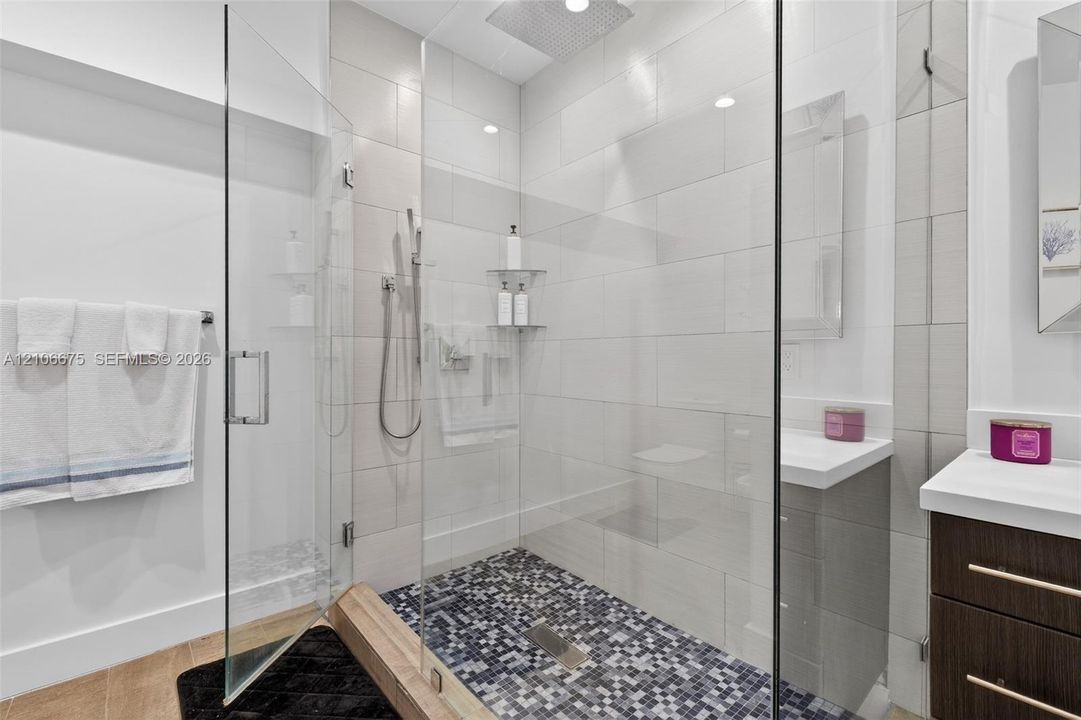 Walk-in shower featuring a glass enclosure, rainfall showerhead, and mosaic tile flooring, complemented by a dark wood-finish vanity with a white countertop