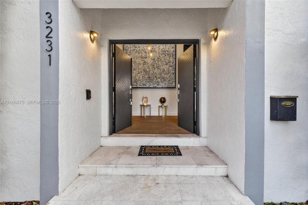 Entryway featuring double-door entrance with chevron-pattern detailing, stone-tiled steps, wall-mounted light fixtures, and exterior building number signage