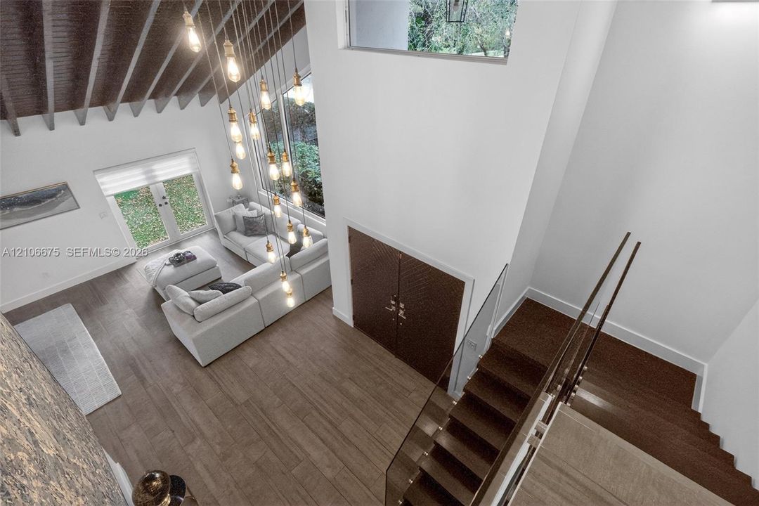 Double-height ceiling with exposed wooden rafters, expansive multi-pane windows, wood-finish flooring, staircase featuring a glass panel railing, and a multi-pendant light fixture