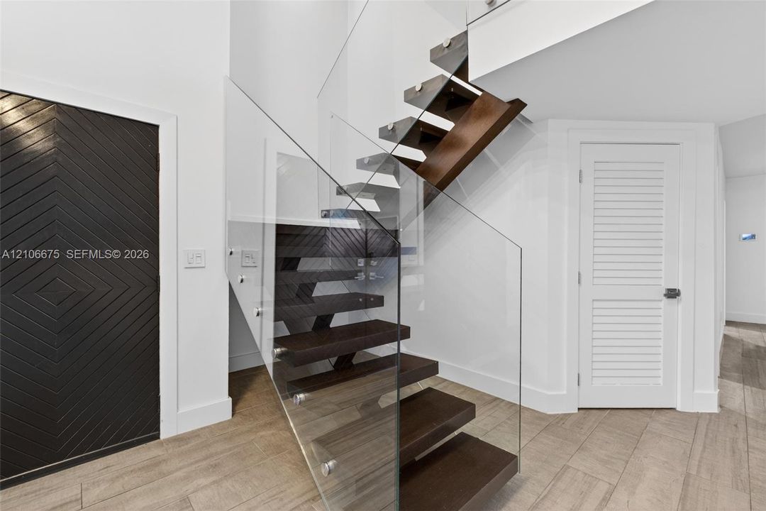 Floating staircase with dark wood-finish treads and frameless glass railing, accented by a geometric patterned door and louvered door, set over wood-finish tile flooring