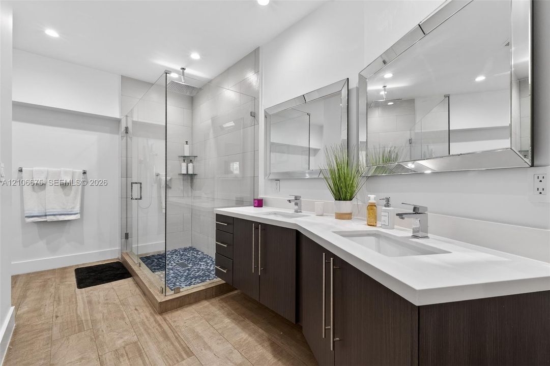 Dual-sink vanity with a white countertop and dark-finish cabinetry, frameless glass shower enclosure with stone tile floor, and large rectangular wall-mounted mirrors
