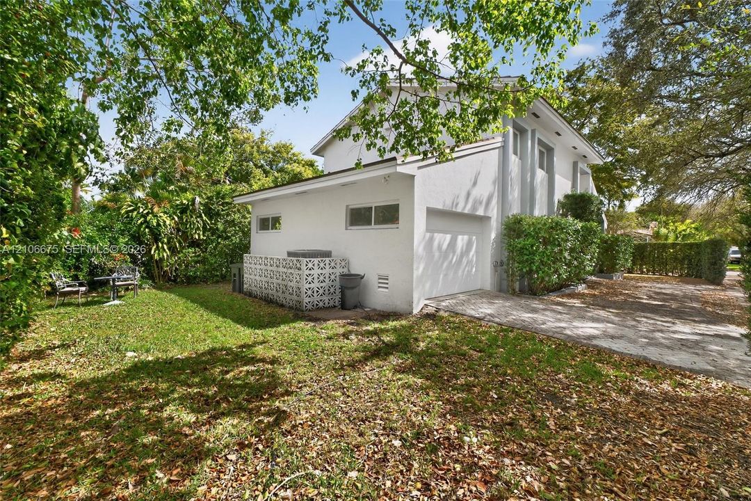 Two-story stucco exterior featuring a built-in privacy screen, attached garage, concrete driveway, and surrounding mature landscaping