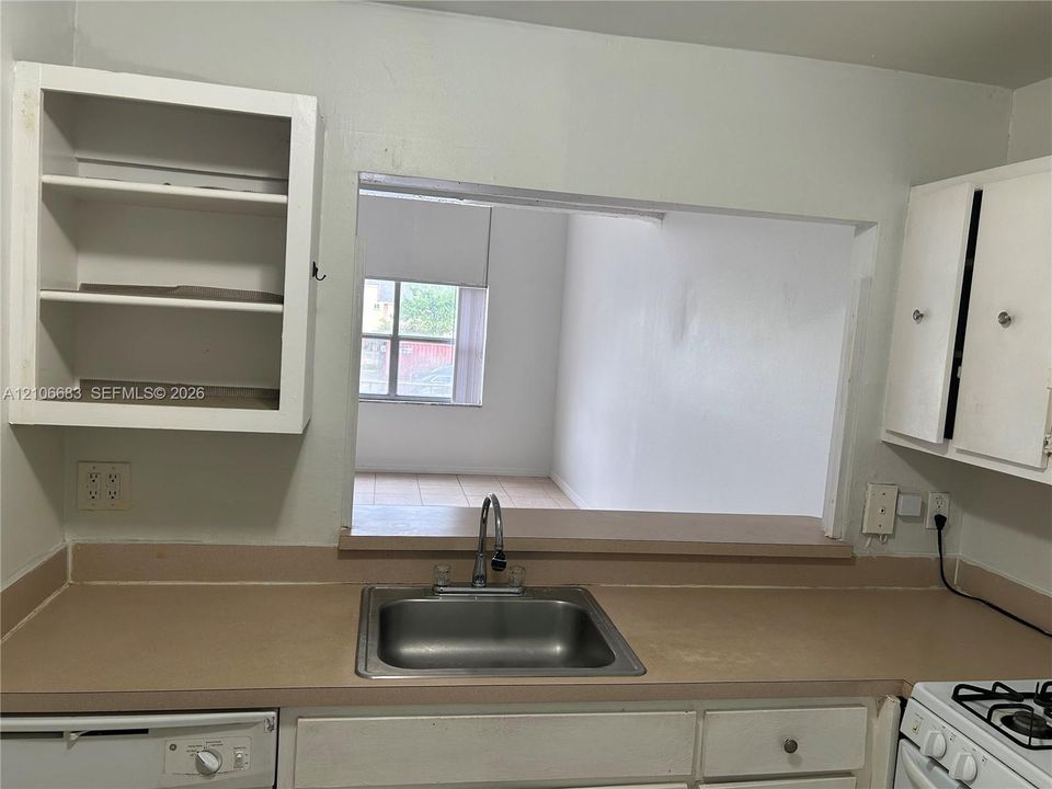 Kitchen featuring a stainless steel drop-in sink, faucet with pull-down sprayer, solid-surface countertop, white cabinetry with open shelving, and a pass-through opening