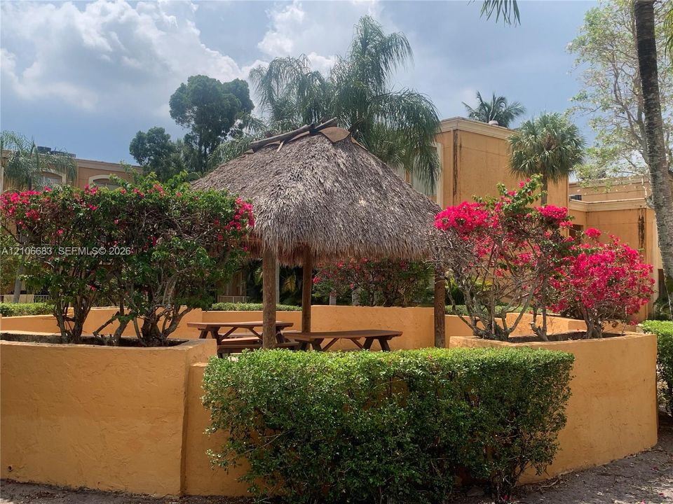 Outdoor palapa structure featuring a thatched roof, surrounded by stucco retaining walls, manicured hedges, and flowering shrubbery