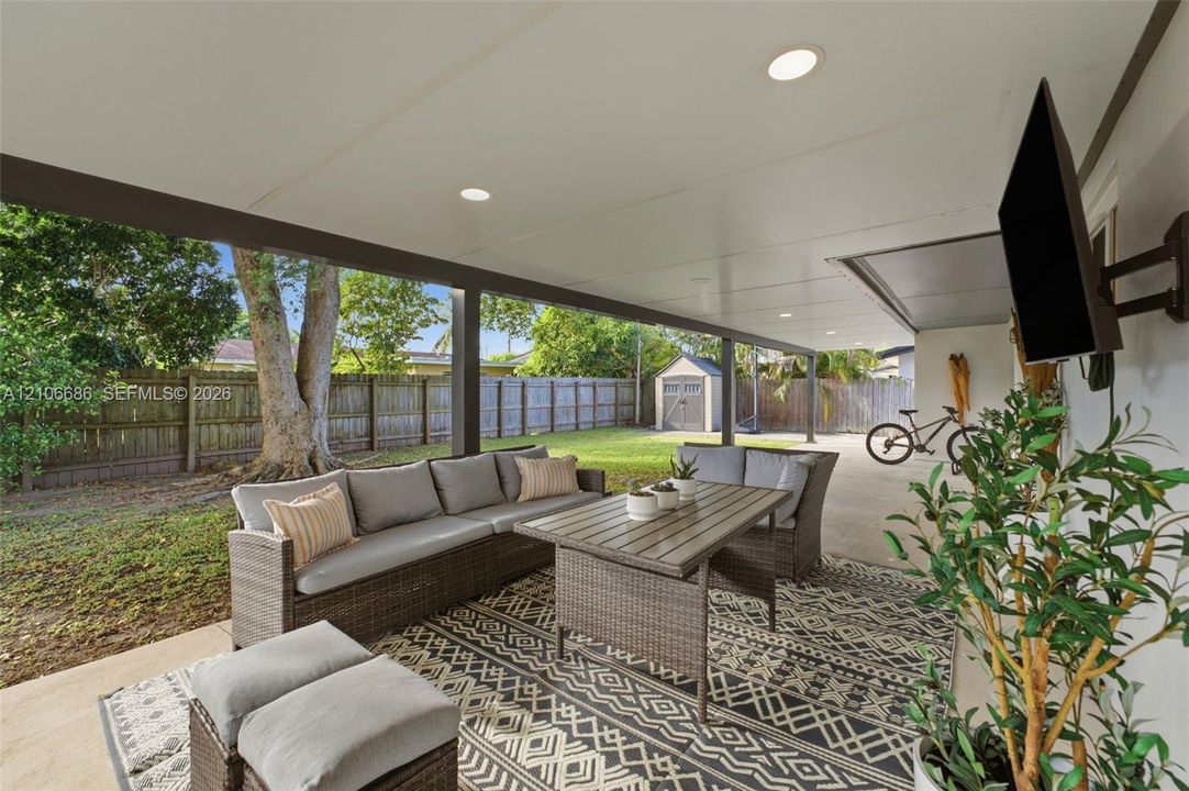 Covered patio featuring a concrete floor, recessed lighting, and a wall-mounted television bracket, overlooking a fenced backyard with a storage shed