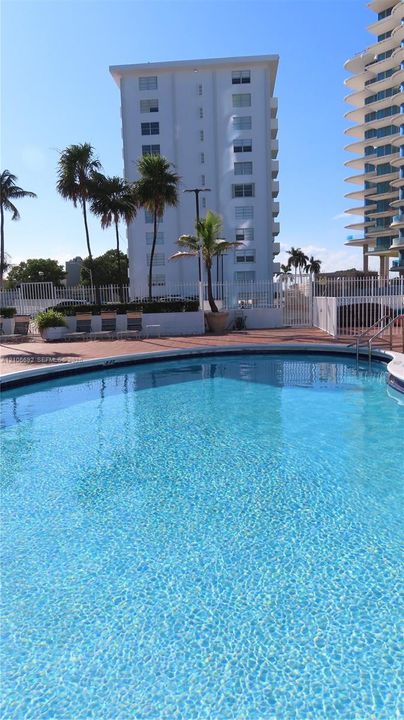 Curved swimming pool with tiled coping, surrounded by a brick-paver deck, white metal perimeter fencing, and mature palm trees