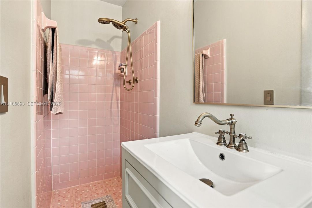 Bathroom featuring square pink tile shower surround, brass-finish handheld shower fixture, rectangular white undermount sink, brass-finish widespread faucet, and large frameless mirror