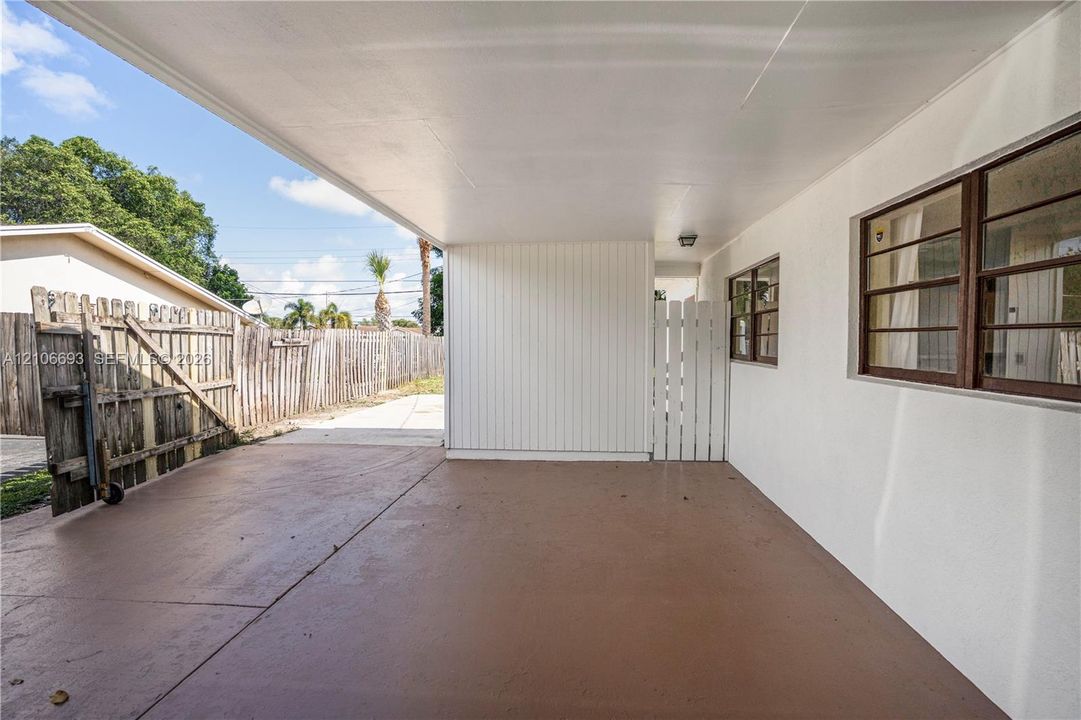 Covered concrete carport featuring white vertical siding, single-hung windows with metal frames, and a white privacy gate