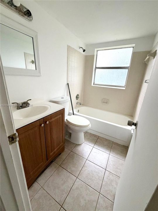 Bathroom featuring a wood-finish vanity with a molded countertop, rectangular wall-mounted mirror, ceramic tile flooring, and a combined tub and shower enclosure with tile surround
