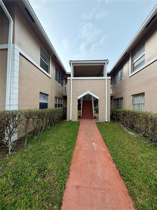 Stucco exterior with multi-level construction, featuring a central covered staircase, concrete walkway, and manicured landscaping