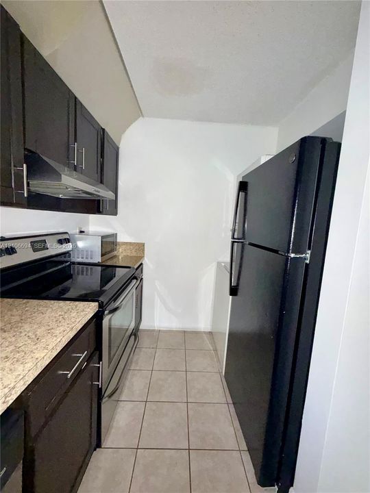 Galley kitchen featuring dark cabinetry, stone-patterned laminate countertops, stainless steel range with overhead hood, and tile flooring