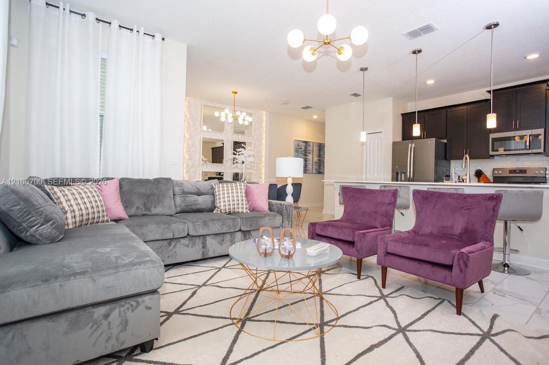 Open-concept area featuring white tile flooring, modern gold-toned multi-globe chandelier lighting, kitchen with dark cabinetry, stainless steel appliances, and a central island with an attached breakfast bar