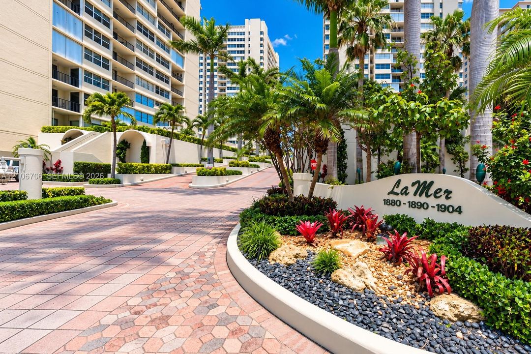 Paver driveway with geometric detailing, manicured tropical landscaping, mature palm trees, multi-story masonry building exterior, and decorative stone accent signage