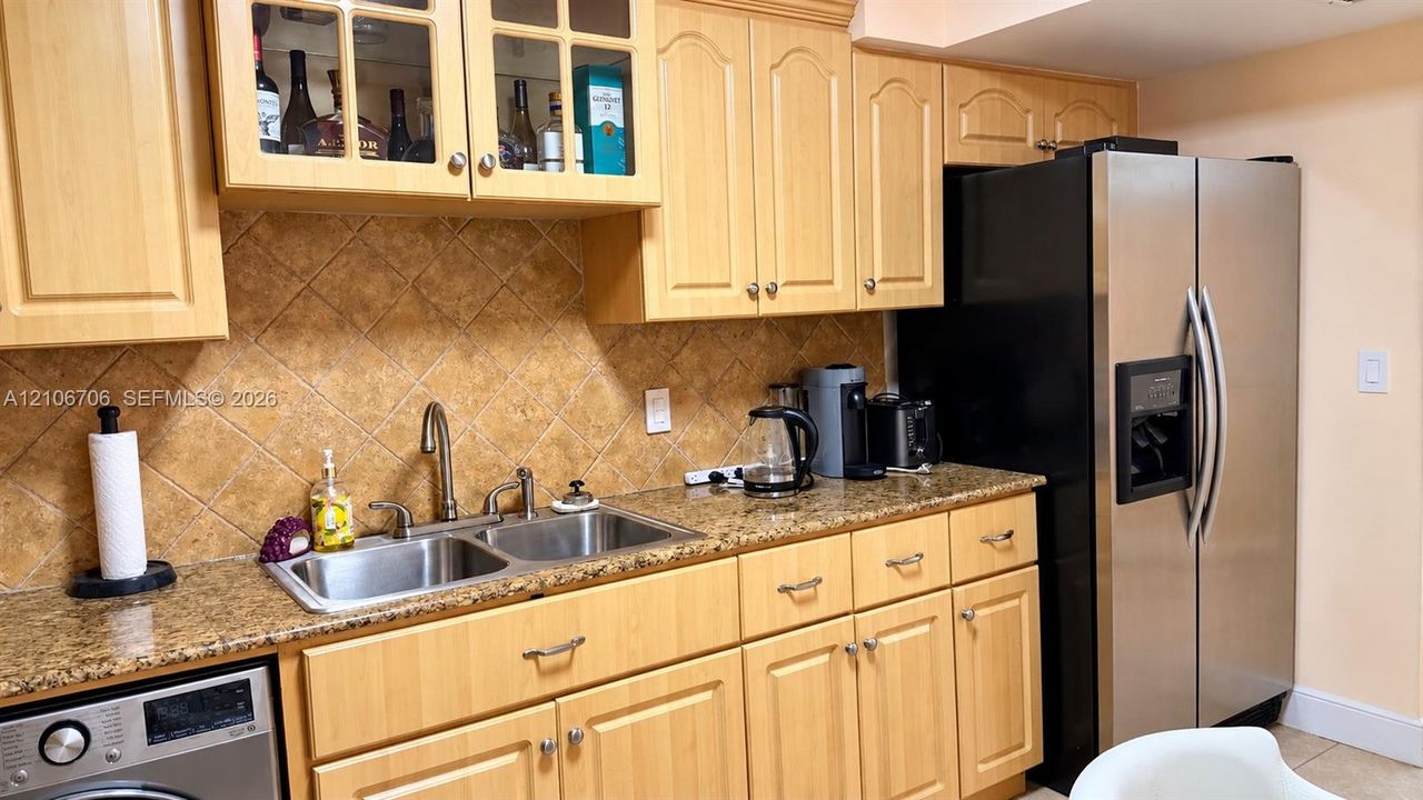 Light-toned wood cabinetry with glass-panel upper doors, stone-finish countertops, diagonal tile backsplash, dual-basin stainless steel sink with gooseneck faucet, and stainless steel refrigerator