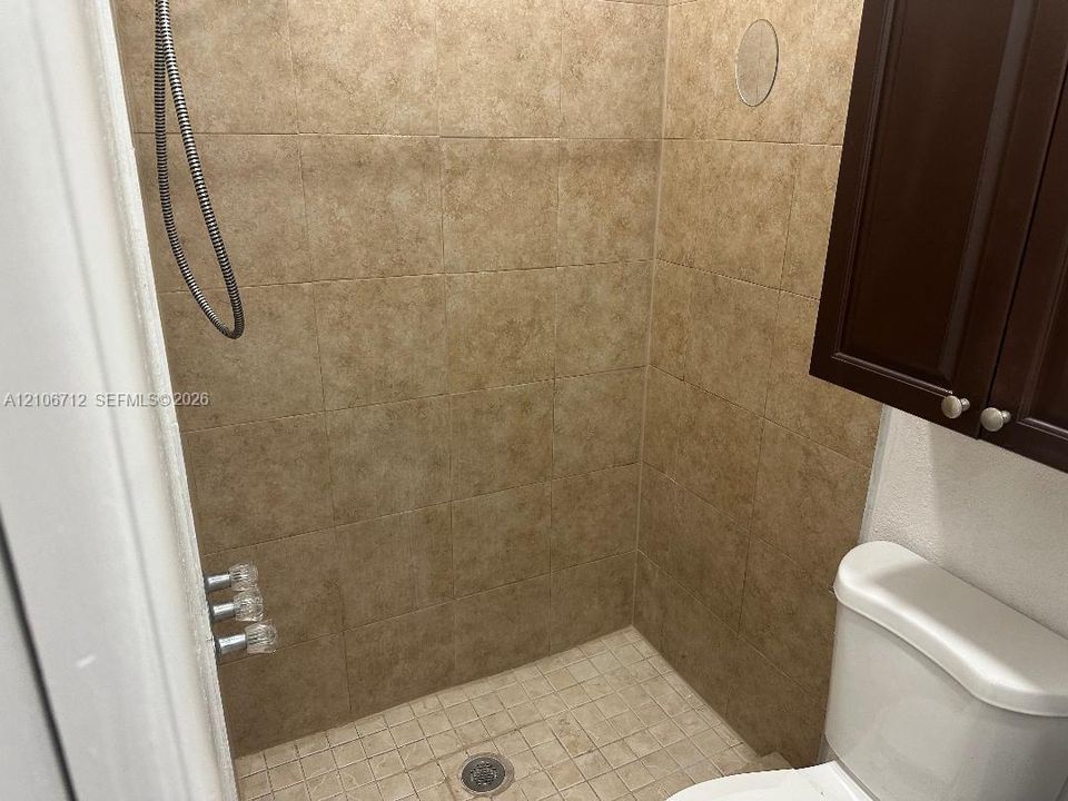 Walk-in shower featuring floor-to-ceiling tile surround, mosaic tile flooring, and metallic fixtures, accompanied by an elevated dark wood-finish cabinet and porcelain commode