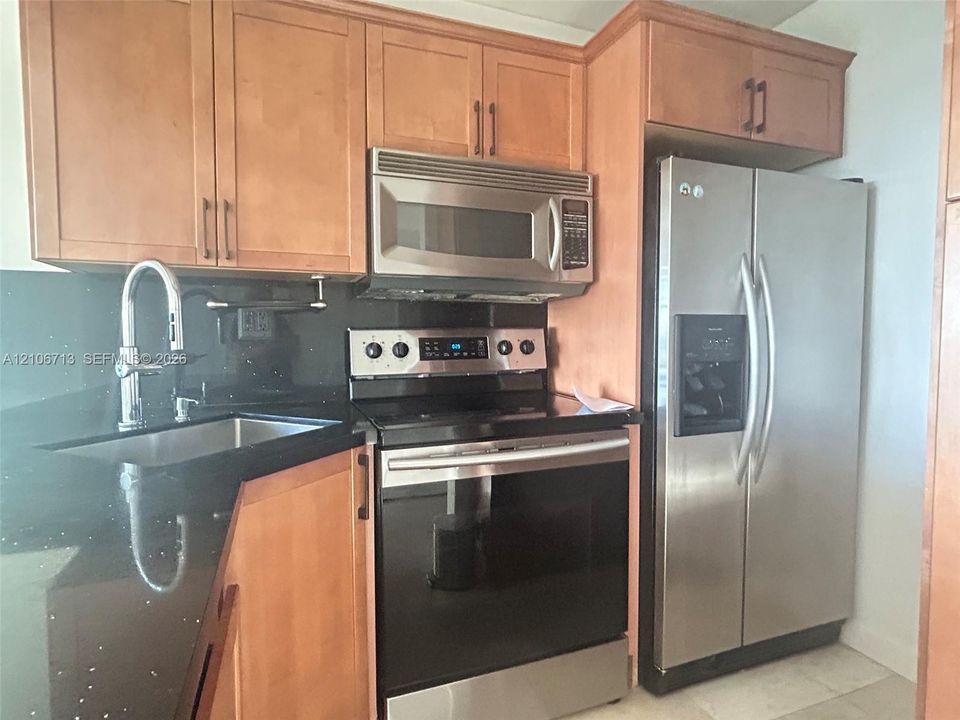 Kitchen featuring wood-finish cabinetry, dark solid-surface countertops, and a suite of stainless steel appliances, including an over-the-range microwave and a side-by-side refrigerator, complemented by a gooseneck faucet