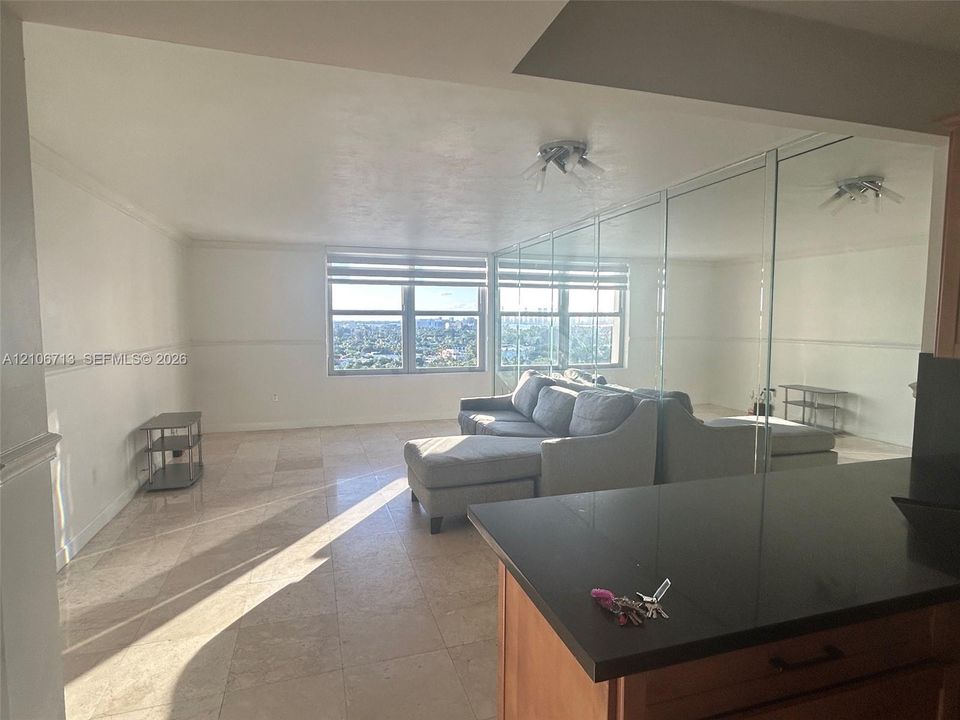 Living area featuring neutral stone flooring, floor-to-ceiling mirrored wall paneling, large windows, and a kitchen peninsula with a dark solid-surface countertop