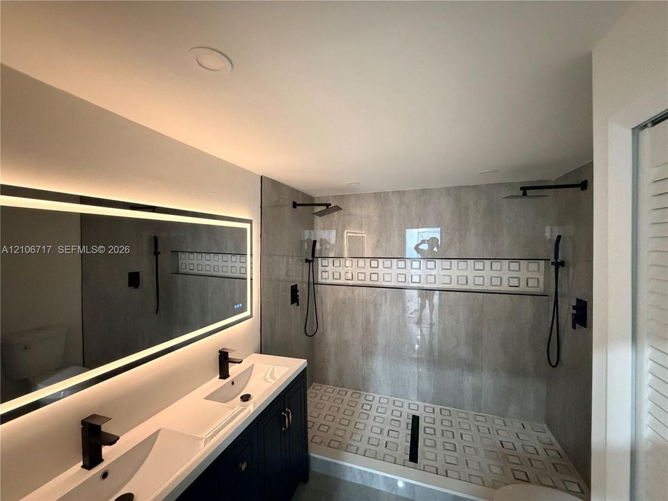 Double vanity featuring integrated sinks, matte black faucets, and a large backlit wall mirror, paired with a walk-in shower showcasing oversized stone-finish wall tile, a decorative square-patterned inlay, and dual shower heads