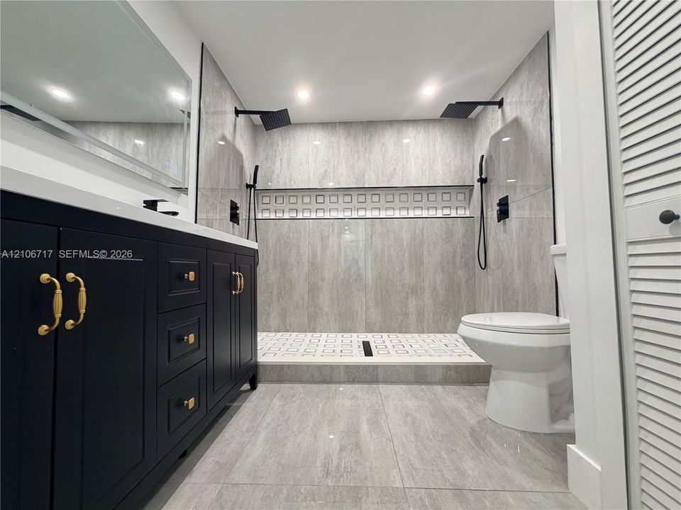 Dark cabinetry with gold-toned hardware, stone-finish flooring, walk-in shower with dual matte black rainfall showerheads, and decorative tile inlay