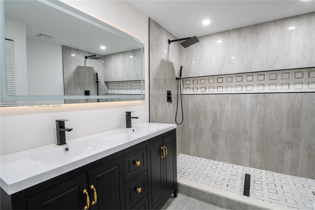 Black vanity with dual undermount sinks, large rectangular mirror with integrated lighting, and walk-in shower featuring matte black rainfall showerhead, textured tile accent wall, and patterned tile flooring
