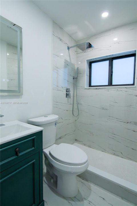 Bathroom featuring a green-toned vanity with a white countertop, a walk-in shower with large-format veined wall tiles, a rainfall showerhead, and a rectangular LED-backlit mirror