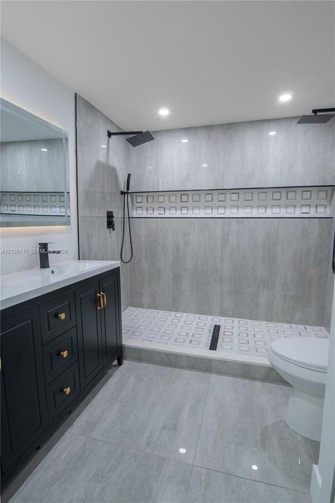 Bathroom featuring a dark wood-finish vanity with gold-tone hardware, white rectangular sink, grey stone-finish tile flooring, and a walk-in shower with dual rain-style showerheads and decorative tile inlay