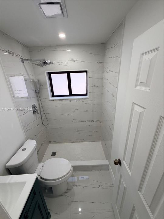 Full-bathroom featuring floor-to-ceiling large-format tile, a walk-in shower with a rainfall showerhead, a modern vanity, and a dual-flush toilet