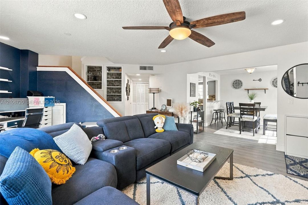 Open-concept living space featuring wood-finish flooring, recessed lighting, a ceiling fan with integrated lighting, and an accent wall with exposed brick detailing