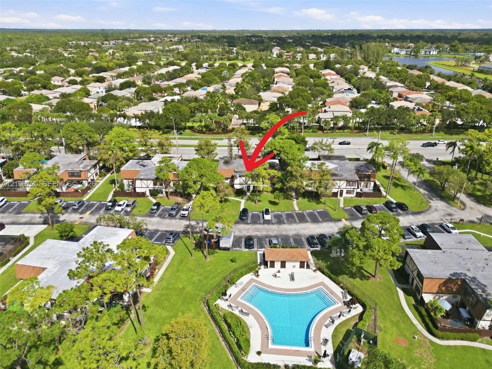 Aerial view of the residential complex featuring multi-unit dwellings, paved surface parking, extensive surrounding greenery, and a community swimming pool