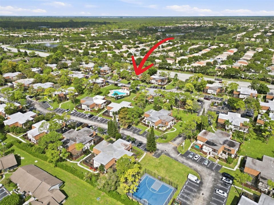 Aerial perspective of residential community featuring a swimming pool, basketball court, paved parking areas, and mature landscaping