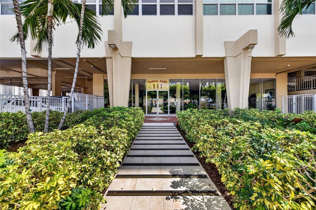 Building entrance featuring structural pillars, floor-to-ceiling glass paneling, a paved walkway with inlaid strips, and manicured landscaping with palm trees