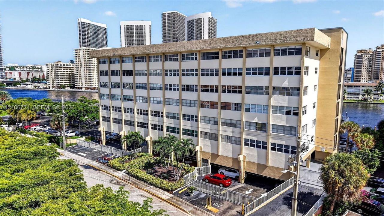 Multi-story residential building featuring a concrete facade, expansive window rows, ground-level covered parking, and perimeter landscaping