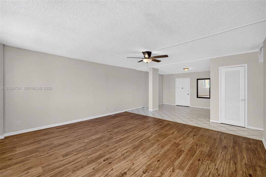 Open interior space featuring wood-finish flooring, neutral wall paint, tiled entryway, ceiling fan with integrated lighting, and a six-panel louvered closet door