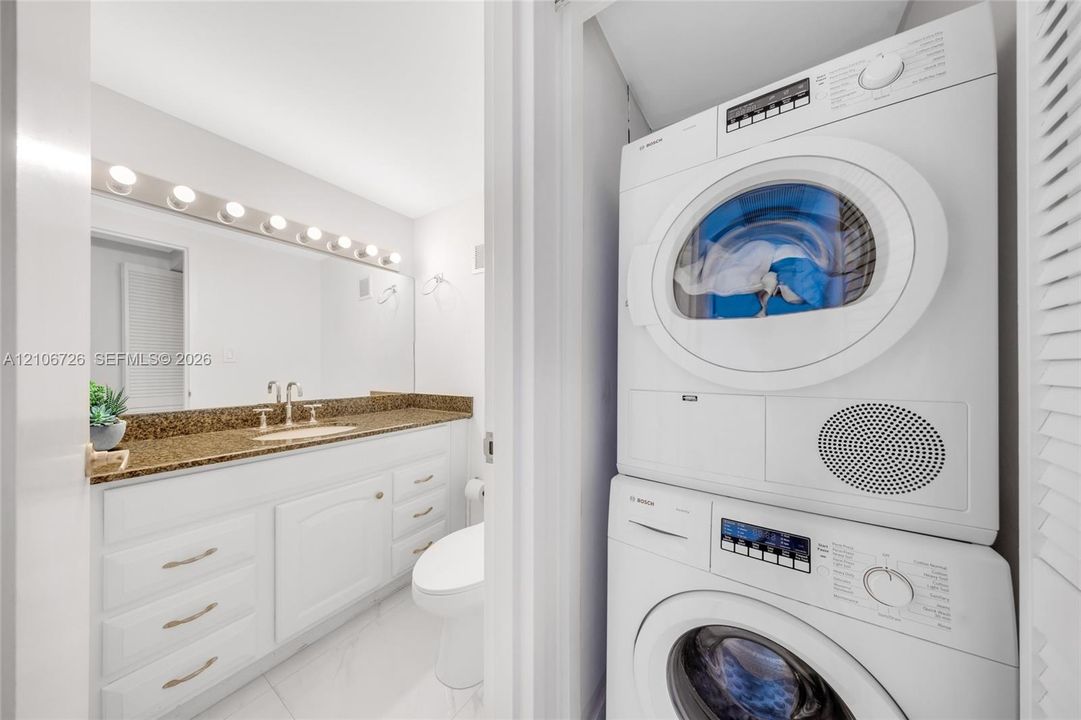 Bathroom featuring white cabinetry, stone-finish countertop, full-width mirror with integrated lighting, and stacked front-load laundry appliances