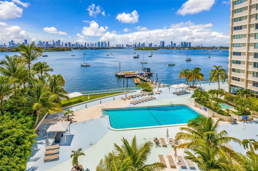 Swimming pool with expansive deck, boat dock, and waterfront positioning featuring palm trees and city skyline backdrop