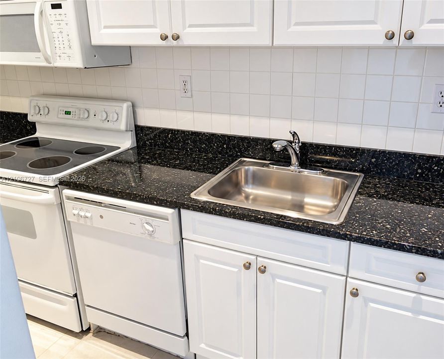 Kitchen featuring white cabinetry, dark speckled stone-surface countertops, a stainless steel undermount sink with a chrome faucet, a white square-tile backsplash, and a white built-in dishwasher