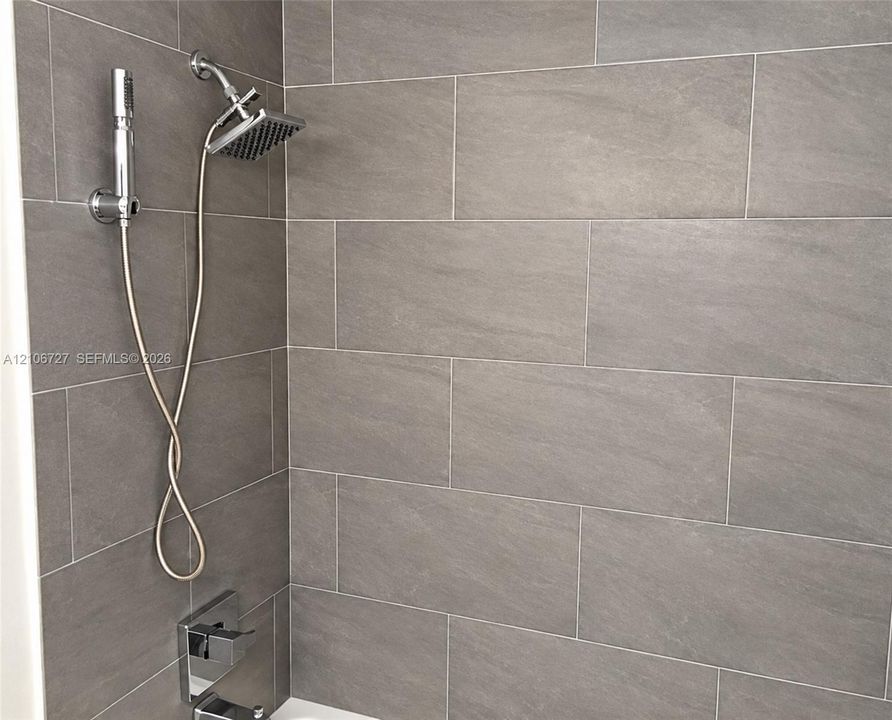 Shower area featuring large-format grey tile wall cladding, a wall-mounted rainfall shower head, a handheld shower wand attachment, and a polished chrome rectangular tub spout